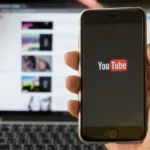 Смартфон з логотипом YouTube у руці перед ноутбуком зі списком відео