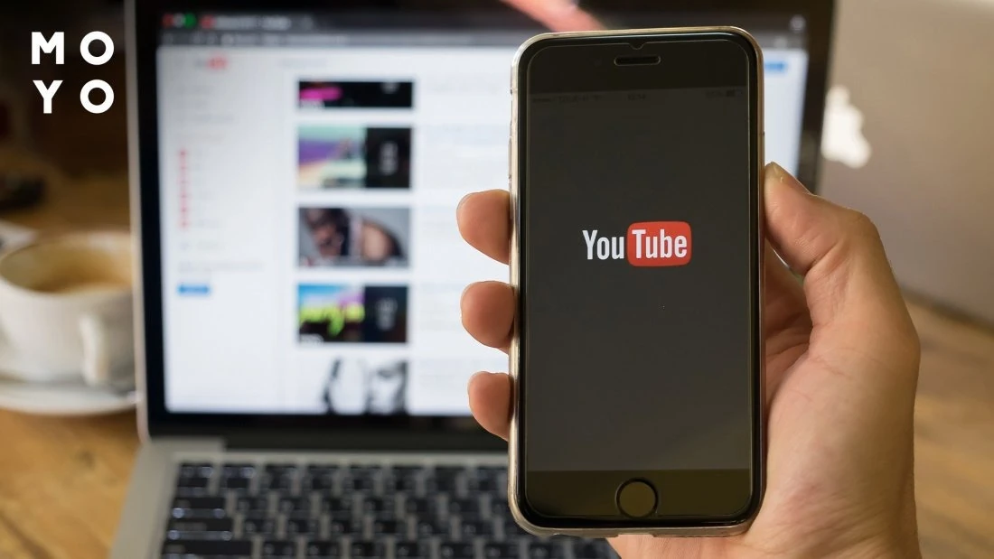 Смартфон з логотипом YouTube у руці перед ноутбуком зі списком відео